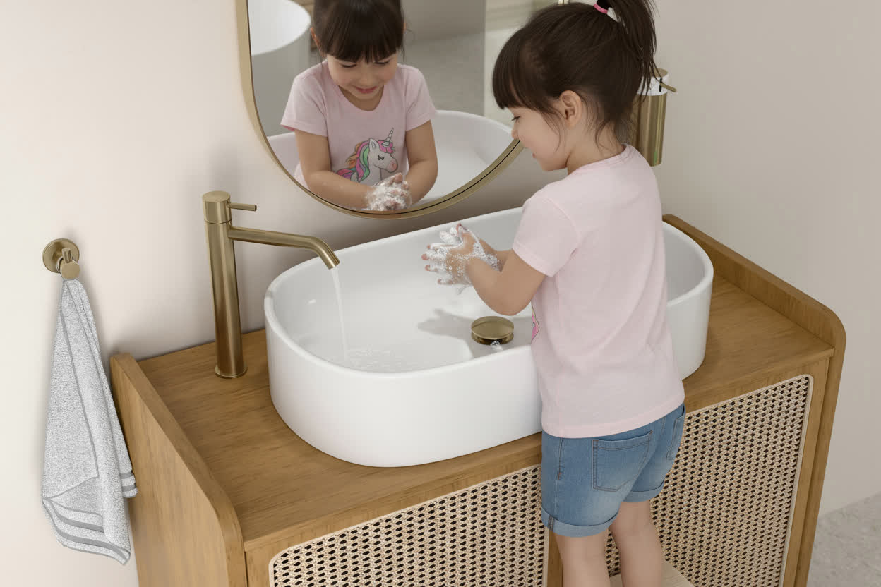 Mała dziewczynka z kucykami myjąca ręce w białej, owalnej umywalce nablatowej, stojącej na drewnianej szafce. W tle widoczne złote akcesoria łazienkowe i lustro, w którym odbija się uśmiechnięta dziewczynka.