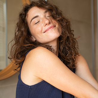 Młoda kobieta z kręconymi włosami i piegami, uśmiechnięta z zamkniętymi oczami, opiera się o lustro w łazience.